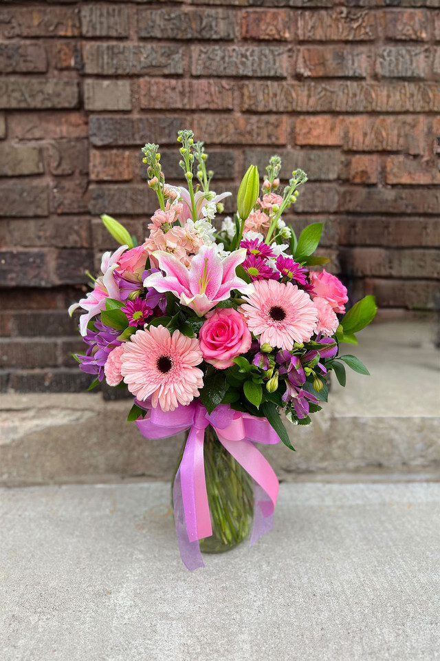 A lush bouquet of pink and purple flowers, including large gerbera daisies, fragrant lilies, roses, and delicate stock, arranged in a glass vase with a flowing pink ribbon.