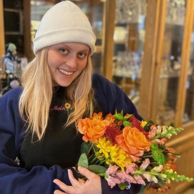 Smiling florist in a beanie and apron holding an autumn bouquet of orange roses, yellow mums, and pink snapdragons.