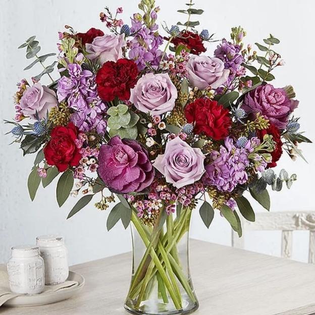 Lush purple and red bouquet with lavender roses, carnations, thistle, and eucalyptus in a tall glass vase.