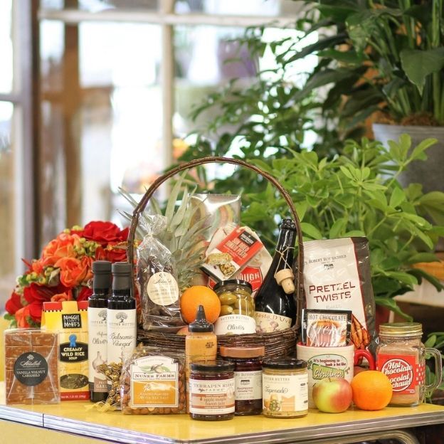 Large gourmet gift basket with snacks, sauces, chocolates, fruit, and wine displayed on a table with greenery behind.