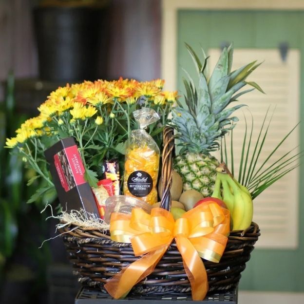 Wicker gift basket filled with fresh fruits, snacks, yellow blooms, and decorated with a large orange and yellow ribbon.