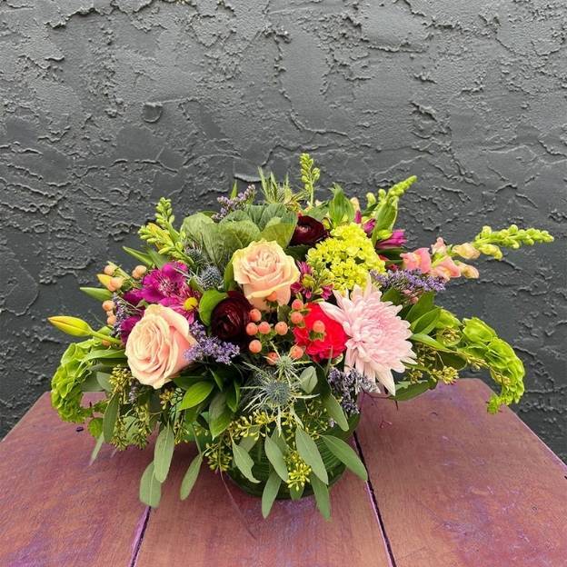 Colorful flower arrangement with roses, chrysanthemums, and greenery in a round vase on a wooden table.