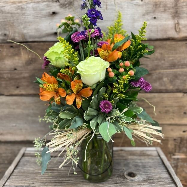 A colorful bouquet in a glass vase with green roses, orange lilies, purple flowers, and greenery on a wooden background.