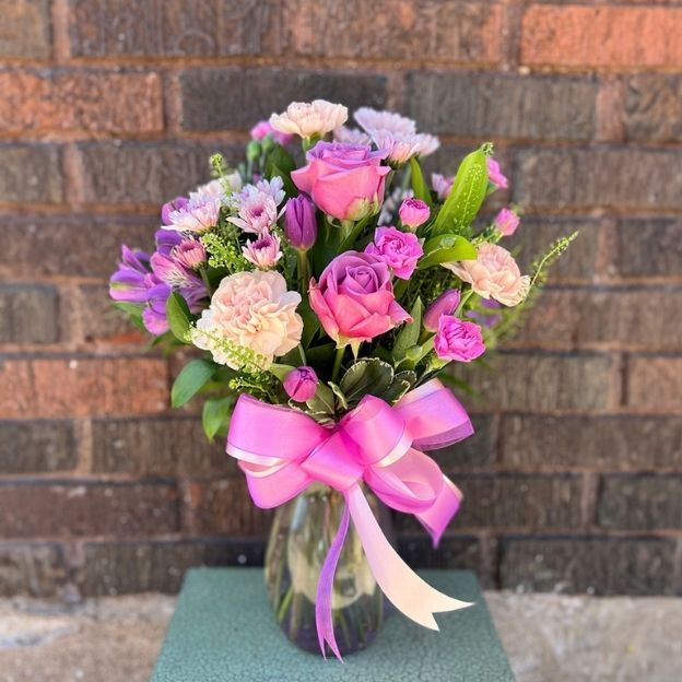 A bouquet of pink and lavender roses, carnations, and daisies in a glass vase tied with a pink and white ribbon.
