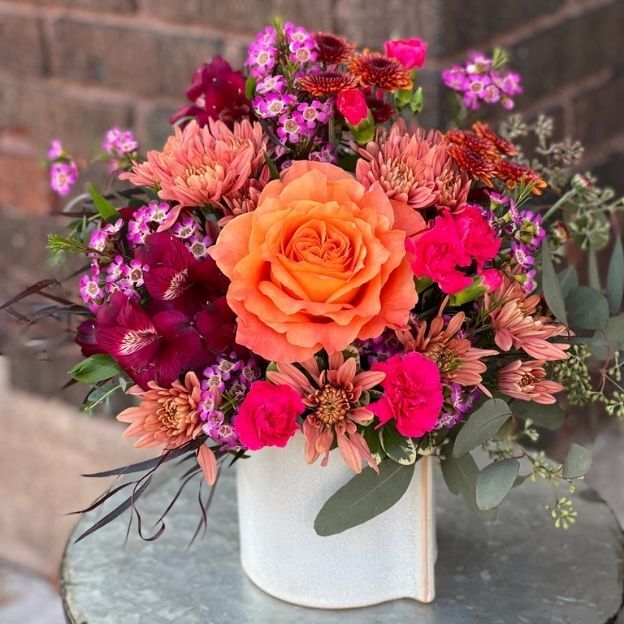 Vibrant flower arrangement with an orange rose, pink carnations, purple blooms, and mums in a white ceramic vase.