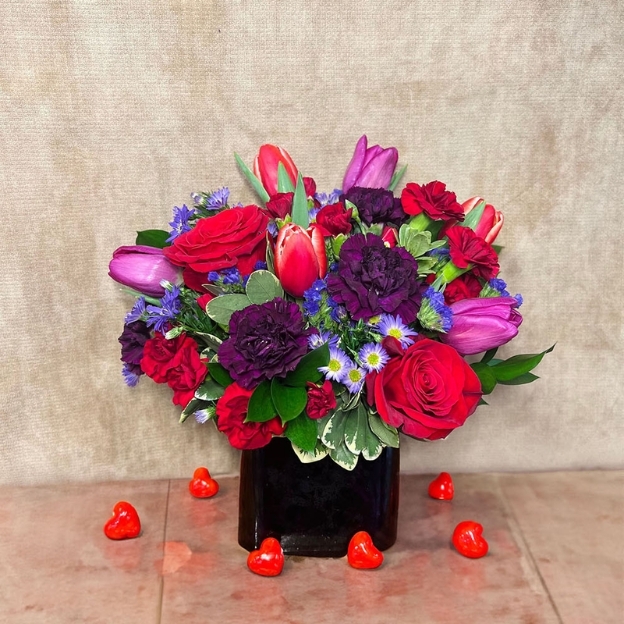 Red roses, purple tulips, carnations, and blue accent flowers in a black vase with heart decorations.