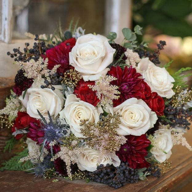 Bridal bouquet with white roses, red dahlias, berries, and greenery arranged in a rustic, natural style.