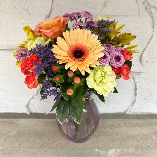 Mixed flower arrangement in a vase featuring peach gerbera, red carnations, purple statice, and green leaves on concrete floor.