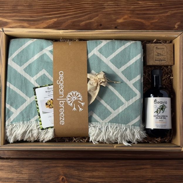 Gift box with a teal patterned towel, extra virgin olive oil, a sachet bag, and a recipe card on a wooden background.