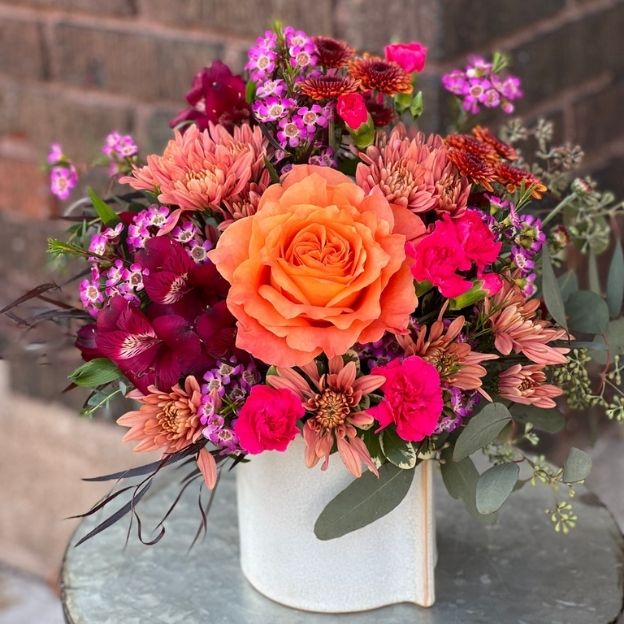Vibrant floral arrangement with an orange rose, pink carnations, and burgundy blooms in a white ceramic vase.