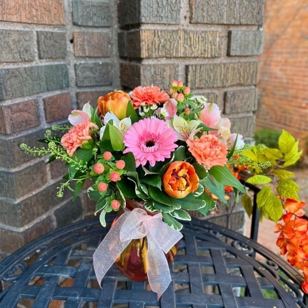 Vibrant bouquet with pink gerbera, orange tulips, and carnations in a vase tied with a sheer bow on a black metal table.