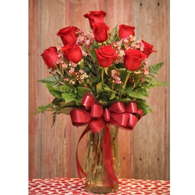 Tall glass vase of red roses with pink waxflower and ferns, tied with a large red bow, on a rustic wood backdrop.