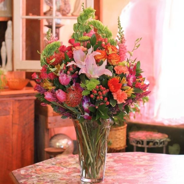 Tall glass vase with pink lily, roses, tulips, carnations, bells of Ireland, and greens on a floral table indoors.