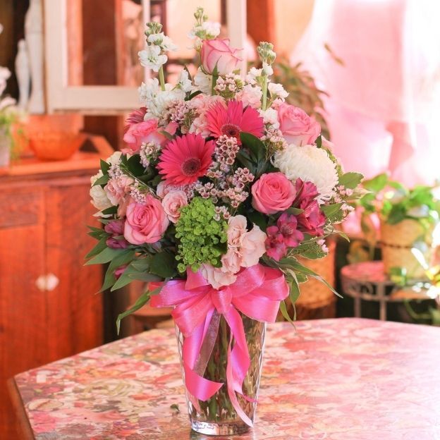 Tall clear vase with pink gerberas, roses, waxflower, white blooms, greenery, and a large pink ribbon bow.