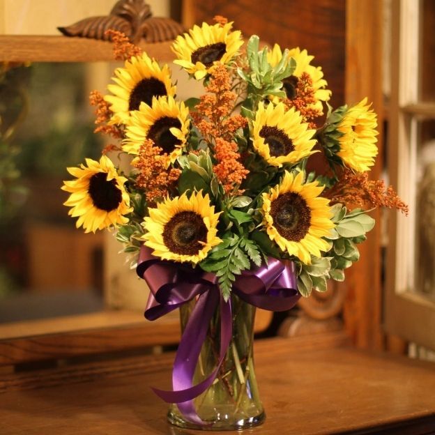 Bright sunflower bouquet with fern and silver-green leaves in a glass vase; purple bow at the base, indoor setting.