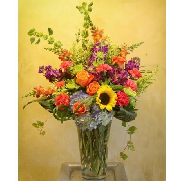 Bright mixed bouquet with sunflower, roses, hydrangea, carnations, and purple blooms in a tall clear vase with greens.