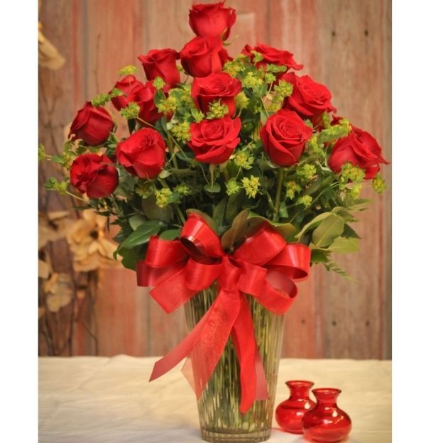 Tall glass vase of long-stem red roses with green filler and a large red bow, set against a rustic wood backdrop.