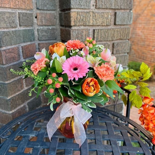 Cheerful spring bouquet with pink gerbera, orange tulips, coral carnations, berries, and greens in a ribboned vase outdoors.
