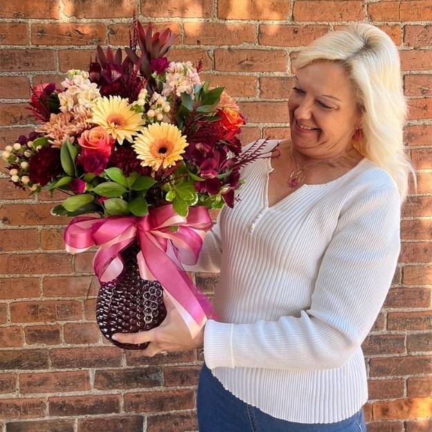 Smiling woman holds a large fall bouquet with gerberas and roses in a textured vase tied with pink ribbon, brick wall behind.