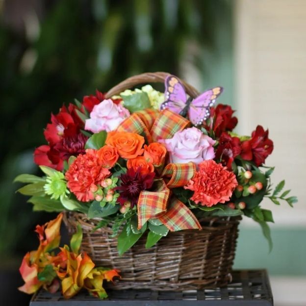 Wicker basket with orange roses, red blooms, lavender roses, berries, plaid bow, and a purple butterfly.