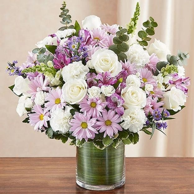 Round bouquet of white roses and carnations with lilac daisies and purple accents in a clear cylinder vase.