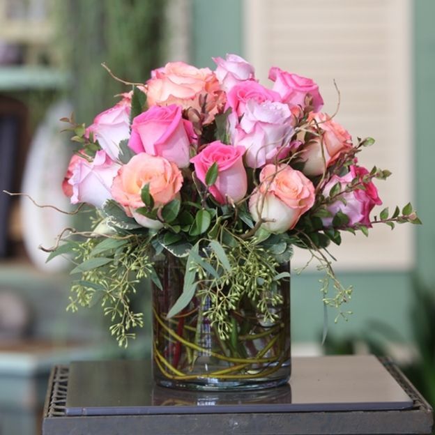 Clustered pink and blush roses with greenery arranged in a glass vase, indoor background softly blurred.