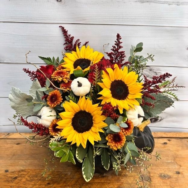 Fall centerpiece with bright sunflowers, burgundy mums, white mini pumpkins, and eucalyptus in a black pumpkin vase.