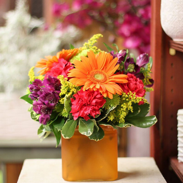Bright mixed bouquet with orange gerberas, magenta carnations, purple blooms, and greenery in a matte orange square vase.