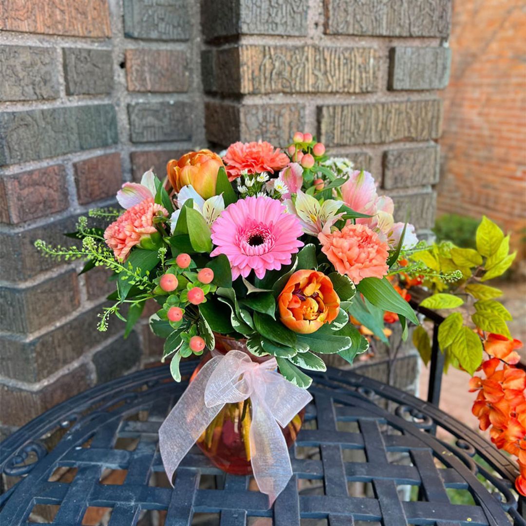 Pink gerbera, orange tulips, coral carnations, and alstroemeria in a ribboned glass vase on a patio table.