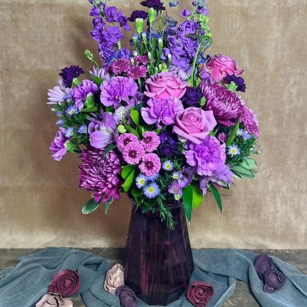 Lavender and purple bouquet with roses, carnations, mums, asters, and alstroemeria in a deep plum glass vase.
