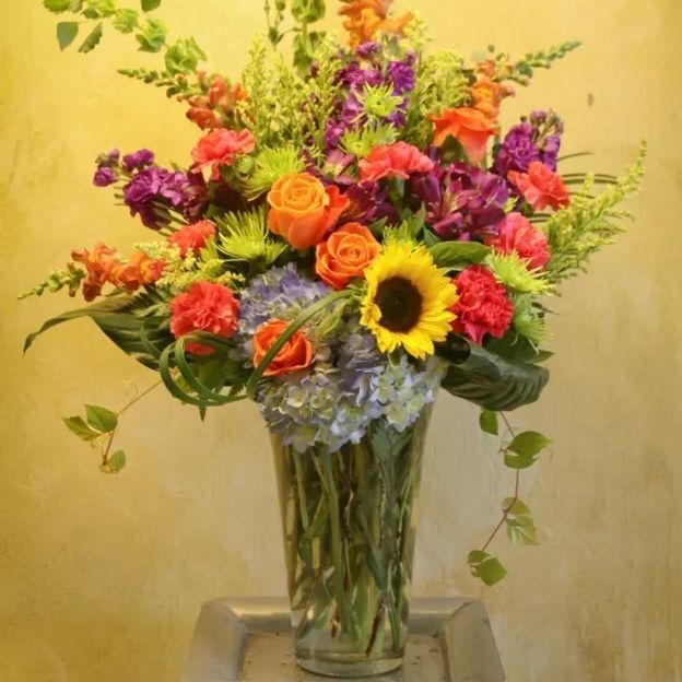 Vibrant arrangement with yellow sunflower, orange roses, purple blooms, blue hydrangea, and greens in a clear vase.