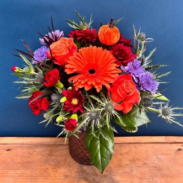 Orange, red, and purple bouquet with silvery thistles and a small pumpkin accent on a wooden table against a blue backdrop.