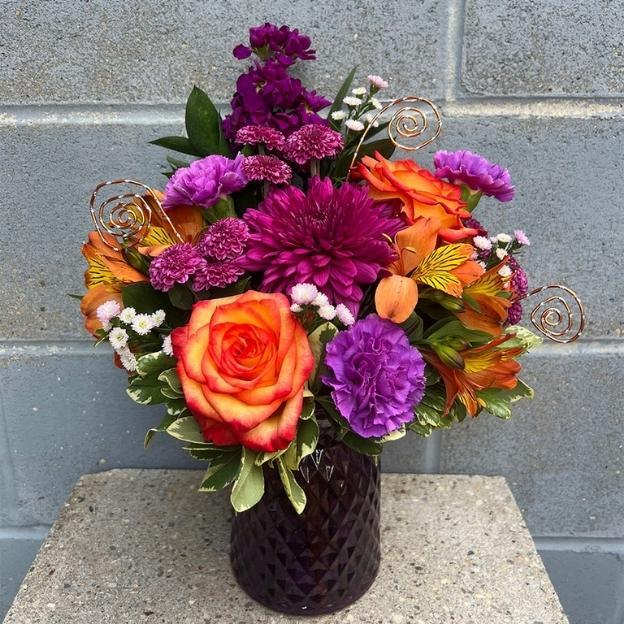 Orange roses, purple carnations, and magenta chrysanthemums with gold wire spirals in a dark purple textured vase.