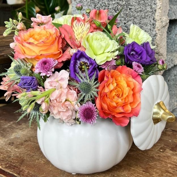 White pumpkin container filled with orange, purple, pink, and pale green flowers, with a matching lid leaning at its side.