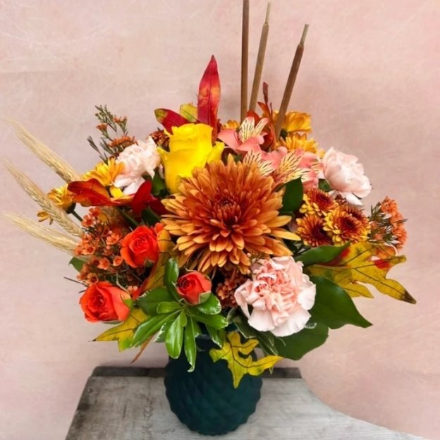Autumn bouquet with bronze blooms, yellow and orange roses, pink carnations, and tall brown reeds in a textured teal vase.
