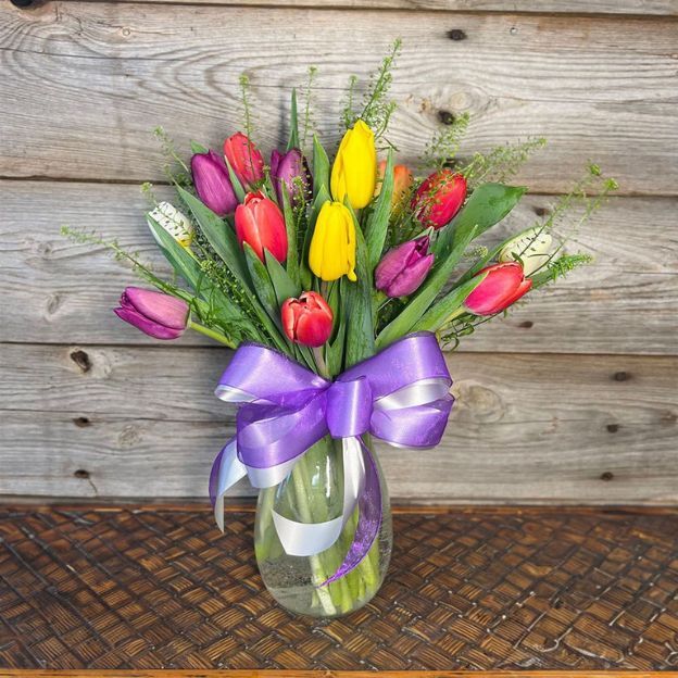 Colorful tulip bouquet in a glass vase with a purple and silver ribbon, set against a wooden background.