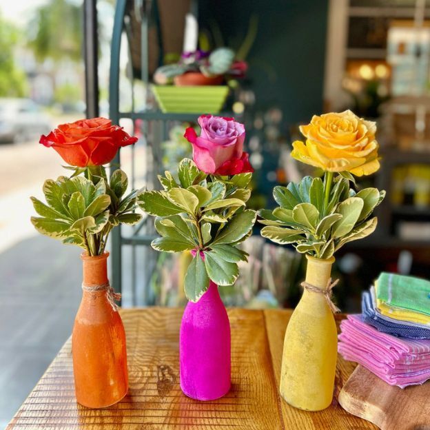 Three single roses in colorful orange, pink, and yellow vases on a wooden table, each with green foliage.